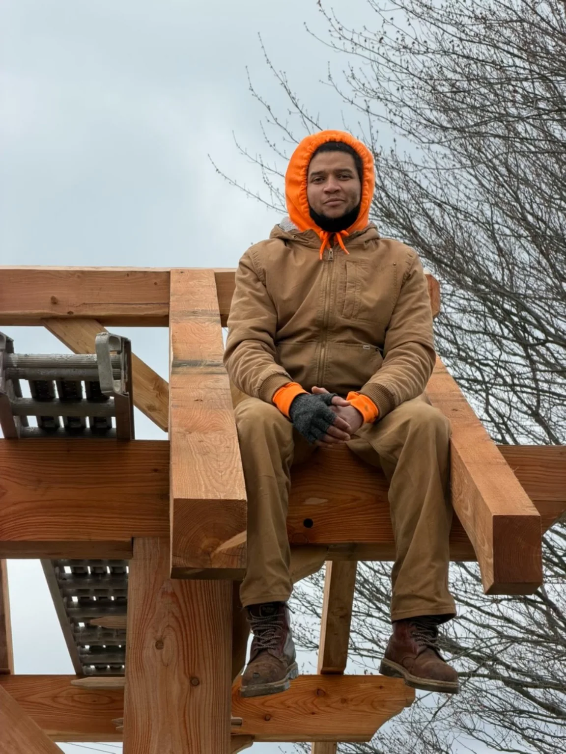 Dom seated on a timber frame during construction, wearing an orange hood and tan jacket.